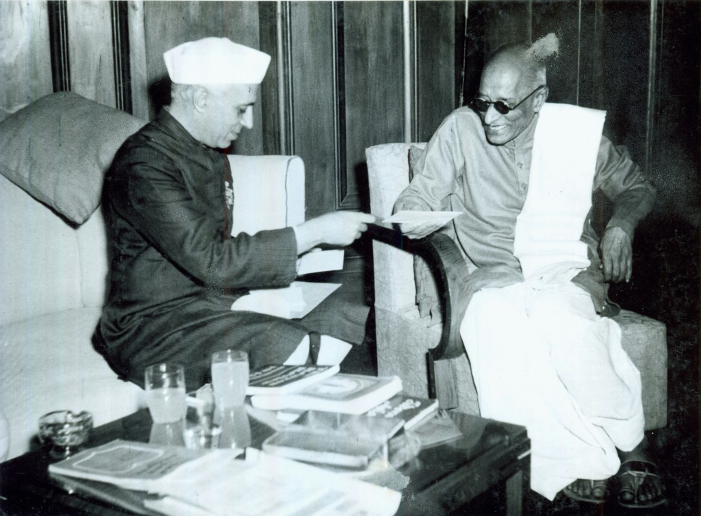 Nehru seated at left passing a paper to Rajaji seated at right, books on the table between them.
