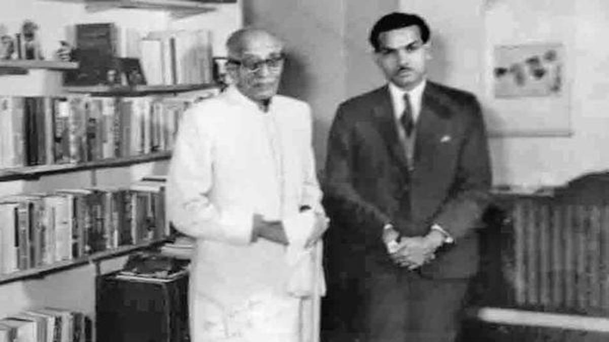 C. Rajagopalachari standing in a room lined with bookshelves beside a younger man in a suit.